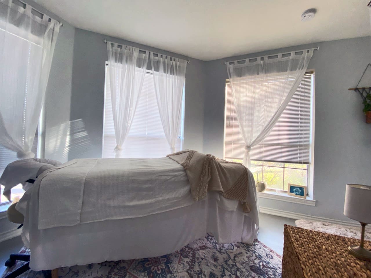 A serene room with soft natural light filtering through sheer white curtains tied at the center. A massage or treatment table covered in white linens and a textured beige blanket sits in the center, placed atop a patterned rug. The space includes a woven side table with a small lamp, a plant on the wall, and a framed piece of artwork on the window ledge, creating a calming and inviting atmosphere.